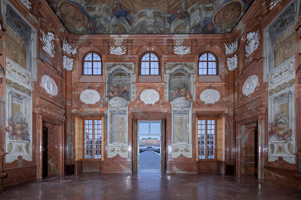 Ornate room with marble walls, large windows, and detailed frescoes. Central door opens to an outside view of a distant building.
