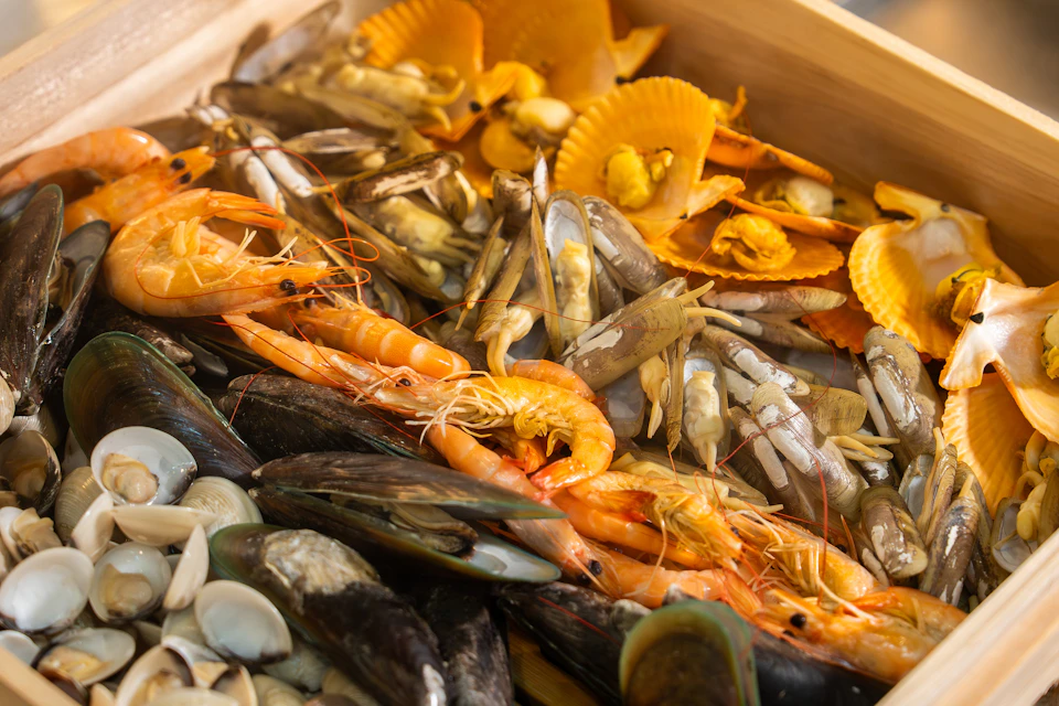 An assortment of fresh seafood including shrimp, mussels, clams, and orange shells displayed on a wooden tray.