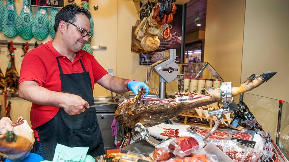 Osoba v červené košili a černé zástěře krájí uzené maso v lahůdkářství s visícím masem a produkty v pozadí.