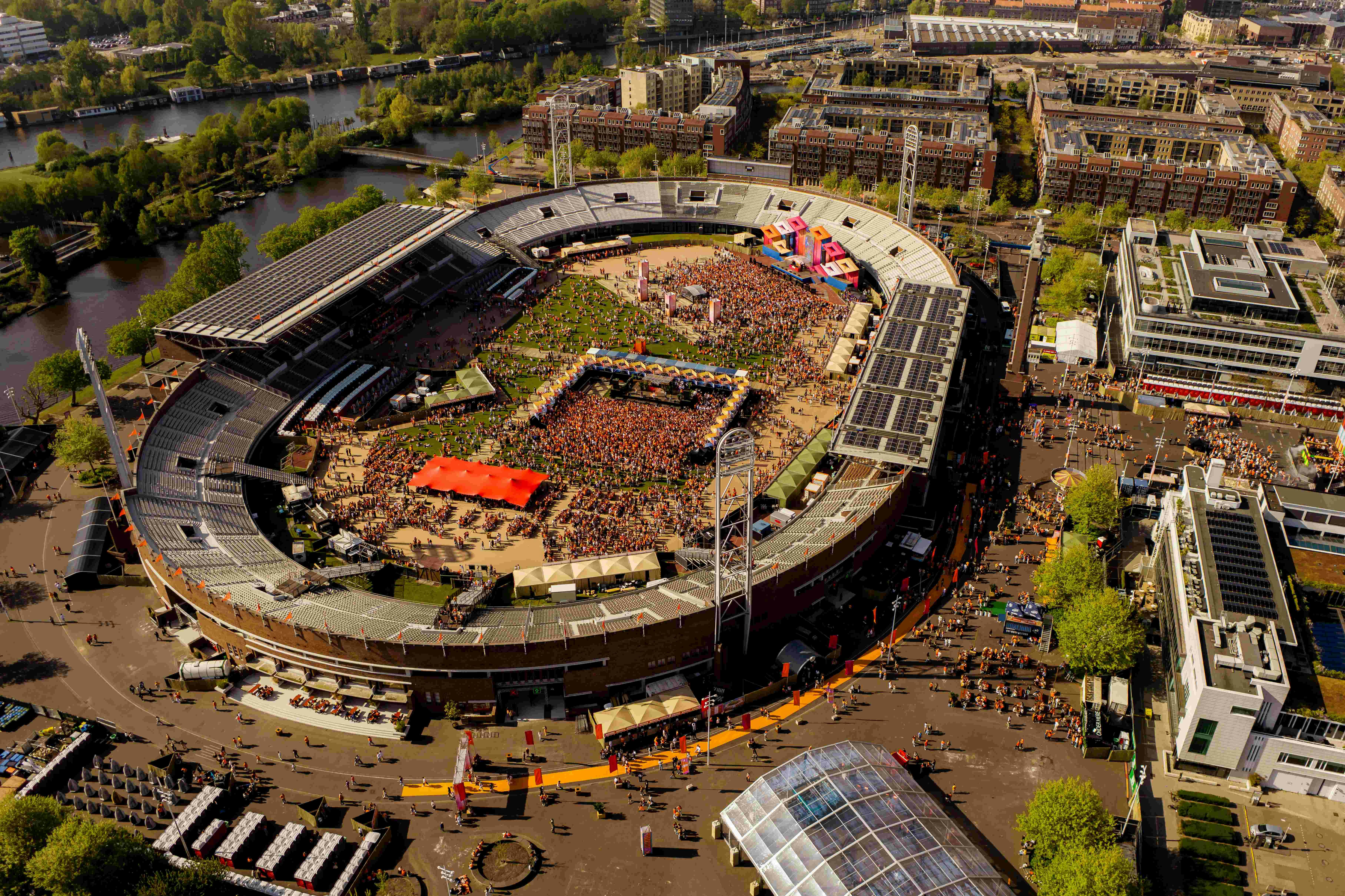 Letecký pohled na velký stadion s přeplněnou venkovní akcí obklopenou městskými budovami a zelení.
