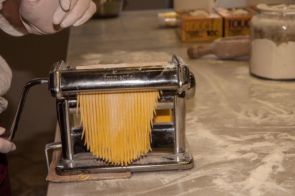 Μια μηχανή ζυμαρικών που εξωθεί φρέσκα φύλλα ζυμαρικών σε έναν αλευρωμένο πάγκο, με ένα χέρι με γάντια και συσκευασμένα ζυμαρικά στο παρασκήνιο.