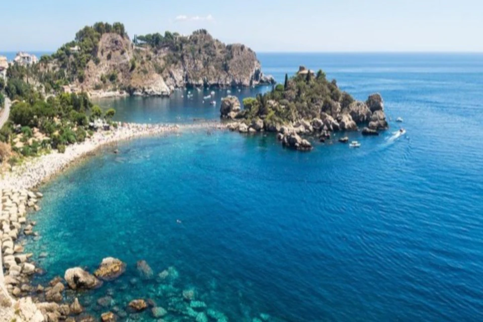 Aerial view of a coastal bay with clear blue water, rocky islands, and a sandy beach alongside a lush, rocky shoreline.