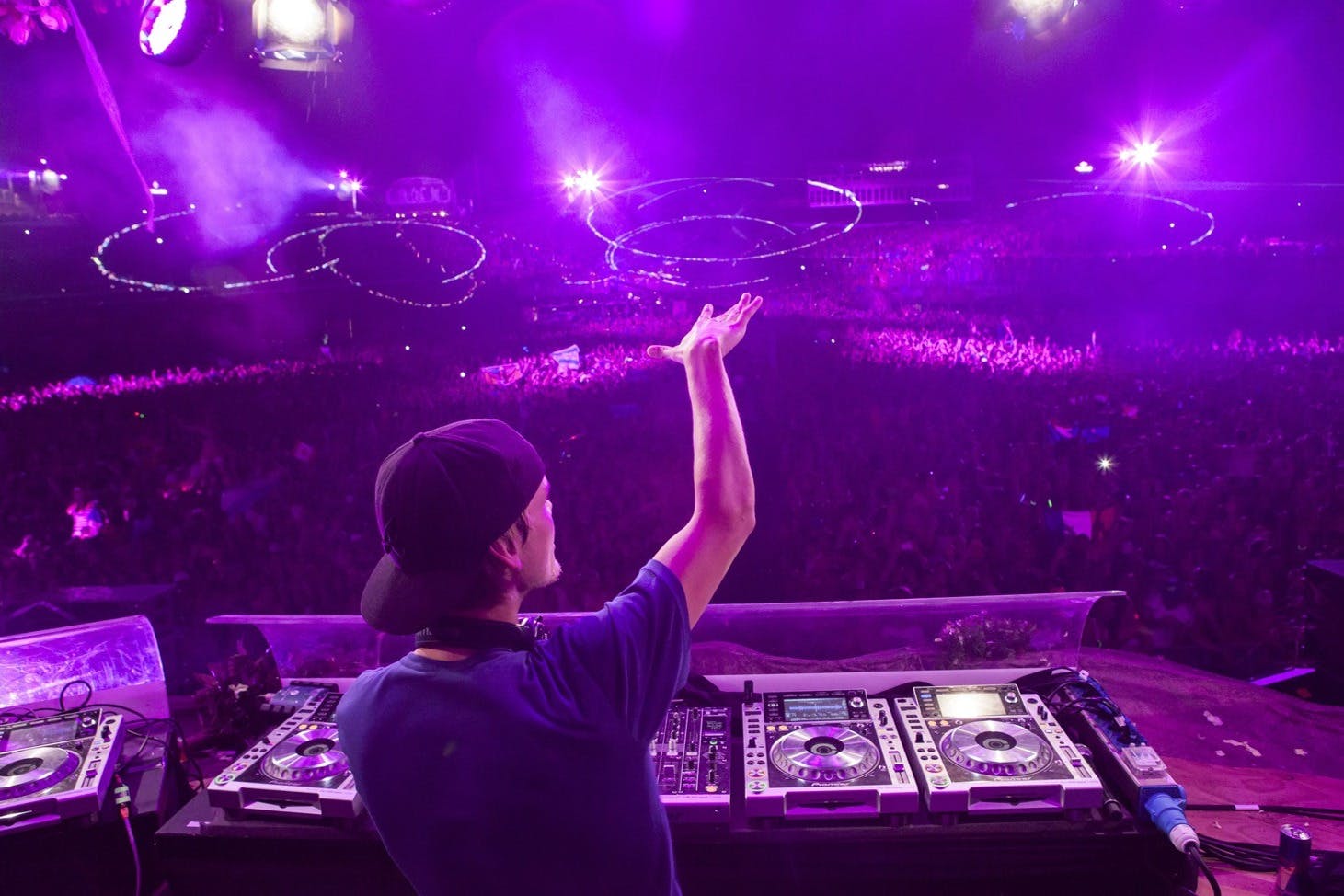 DJ performing on stage at a large festival, with purple lighting and a crowd of fans extending into the distance.
