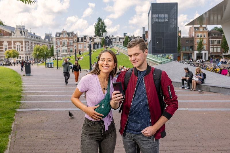 Una donna e un uomo sorridenti guardano insieme uno smartphone in una strada cittadina con erba verde ed edifici sullo sfondo.