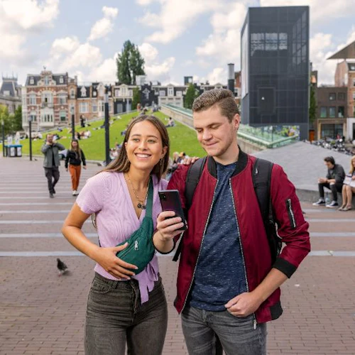 En leende kvinna och man tittar på en smartphone tillsammans på en stadsgata med grönt gräs och byggnader i bakgrunden.