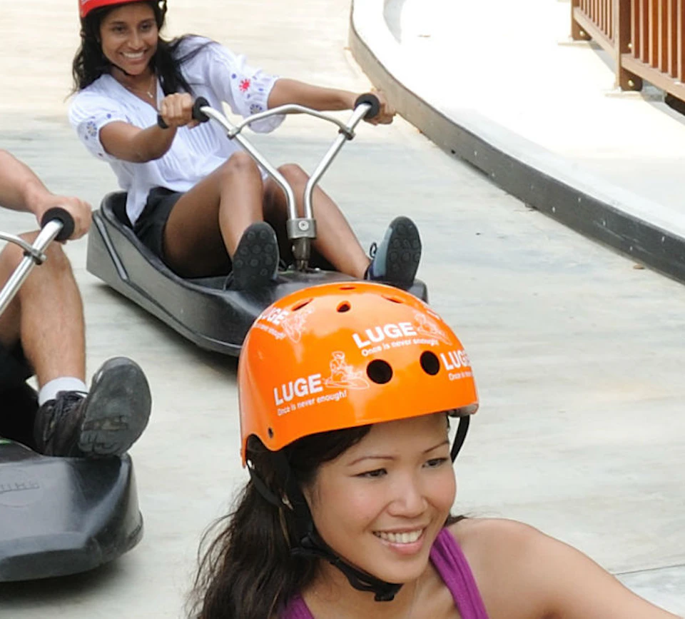 Four people in helmets riding on small, black, sled-like go-karts on a paved track surrounded by greenery.