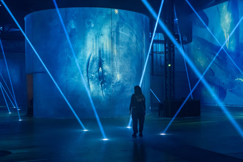 A person stands in a dimly lit space with blue lights and beams, facing a large screen displaying a mysterious eye-like image.