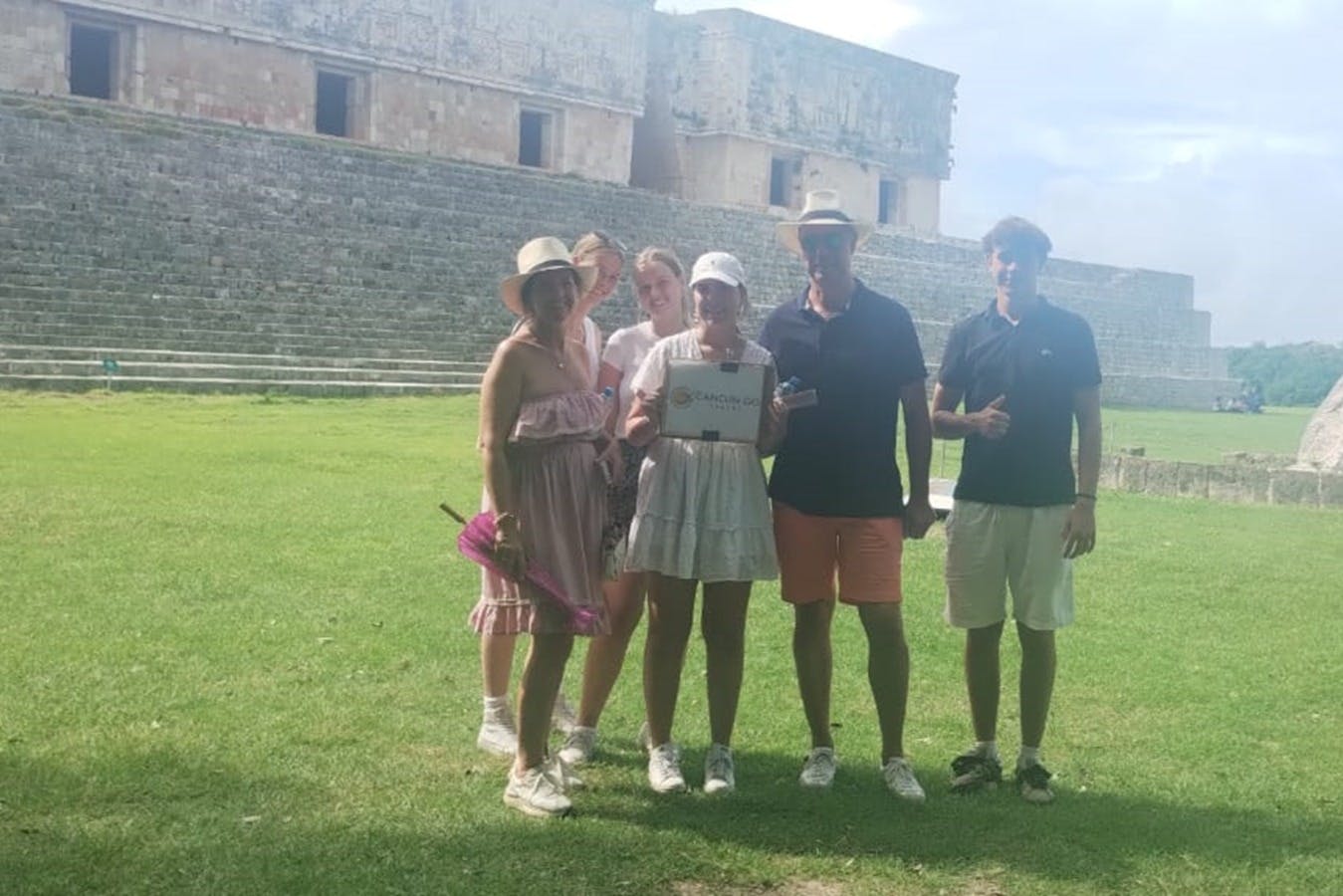 Un groupe de cinq personnes se tient sur l'herbe devant une ancienne structure en pierre avec des sculptures et des escaliers complexes.