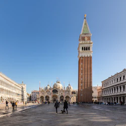 Stort öppet torg med historiska byggnader, ett högt klocktorn och en katedral. Folk går runt under en klarblå himmel.