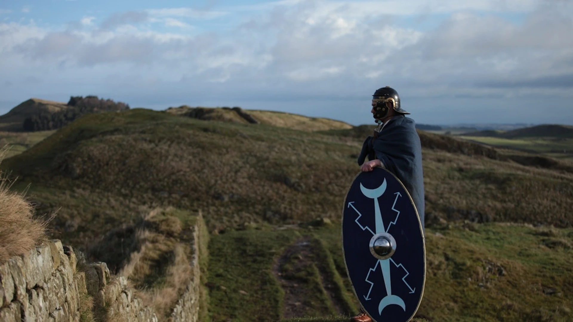 A person wearing a helmet and cloak holds a shield while standing on a grassy hilltop under a cloudy sky.