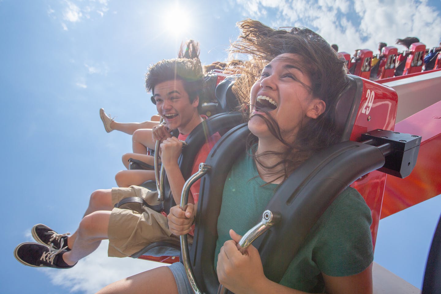 Montagne russe con binari e corridori rossi, circondate da alberi verdi, sotto un cielo blu con nuvole sparse.