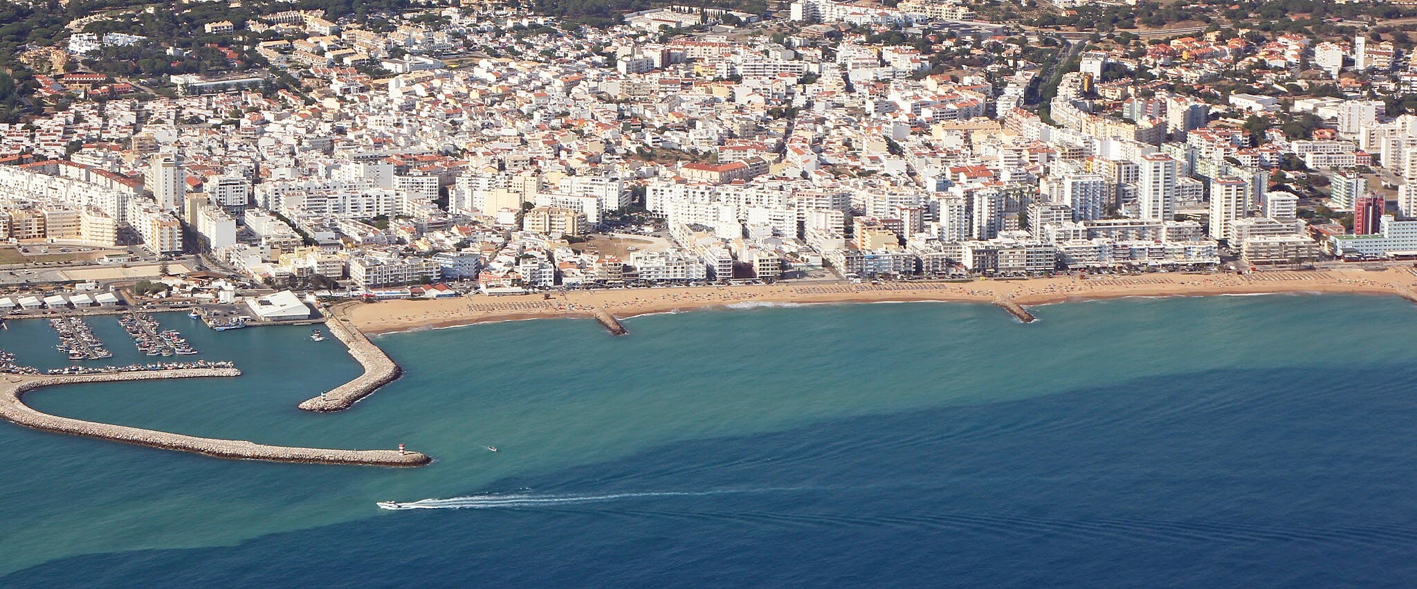 Vista aérea de una ciudad costera con numerosos edificios blancos, una playa de arena, un puerto deportivo y varios barcos en el agua.