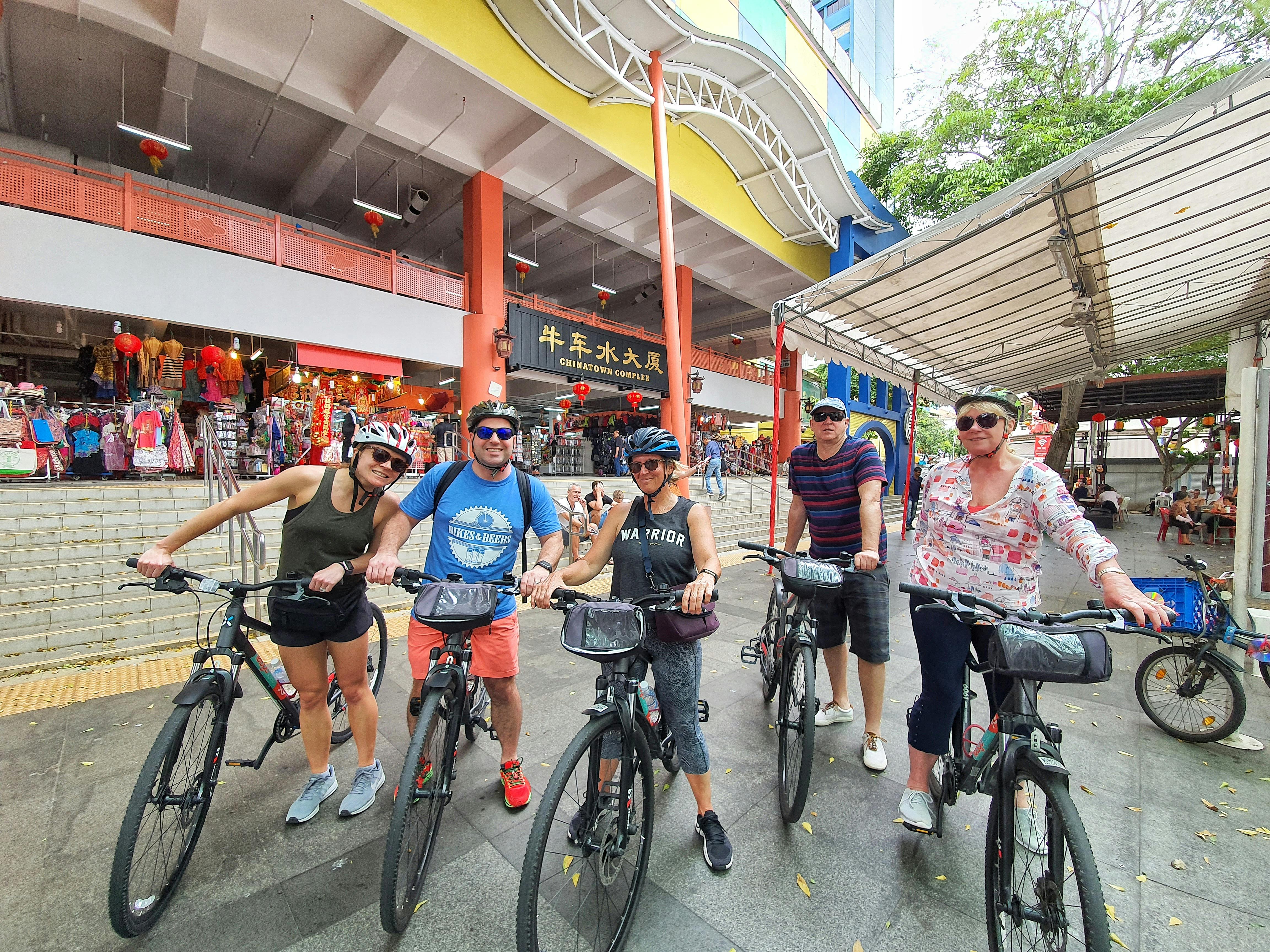 Fire personer med cykler poserer foran en farverig kommerciel bygning med butikker og skiltning, smilende og iført hjelme.