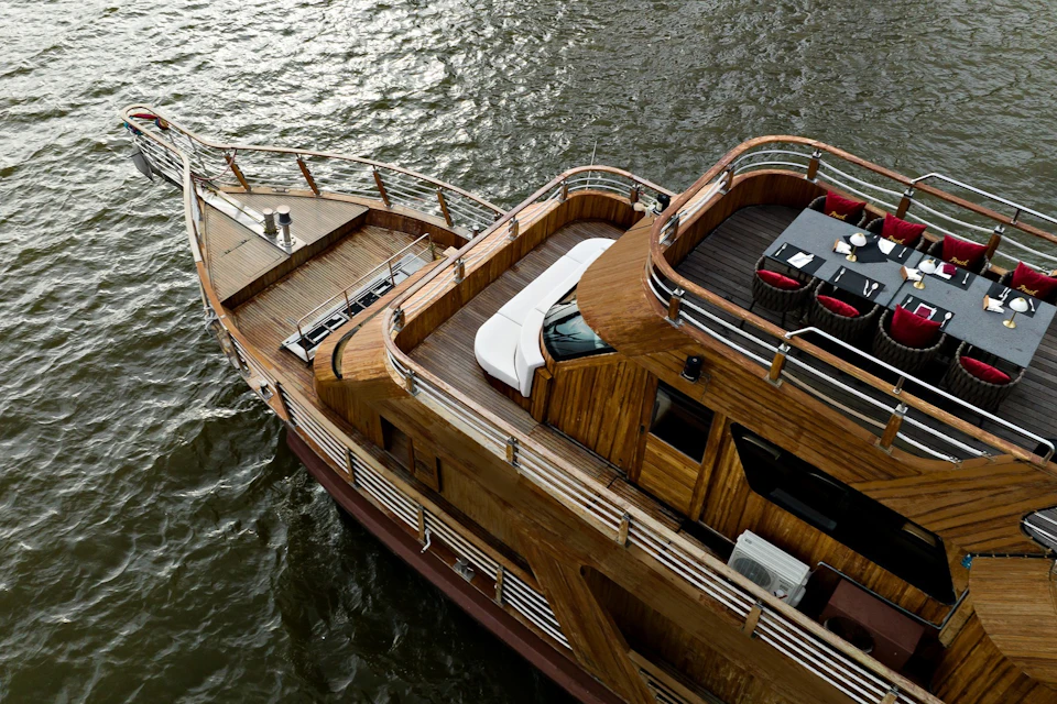 A wooden yacht on the water, featuring decks with railings, a dining area with a table and red chairs, and outdoor seating.