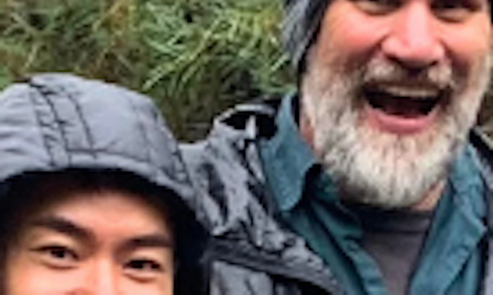 Three people in outdoor attire stand on a wooden path over a stream, smiling and giving thumbs up, with a rocky, forested background.