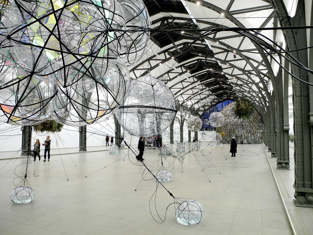 Umělecká instalace v prostorné hale s velkými průhlednými kulovitými konstrukcemi spojenými černými dráty.