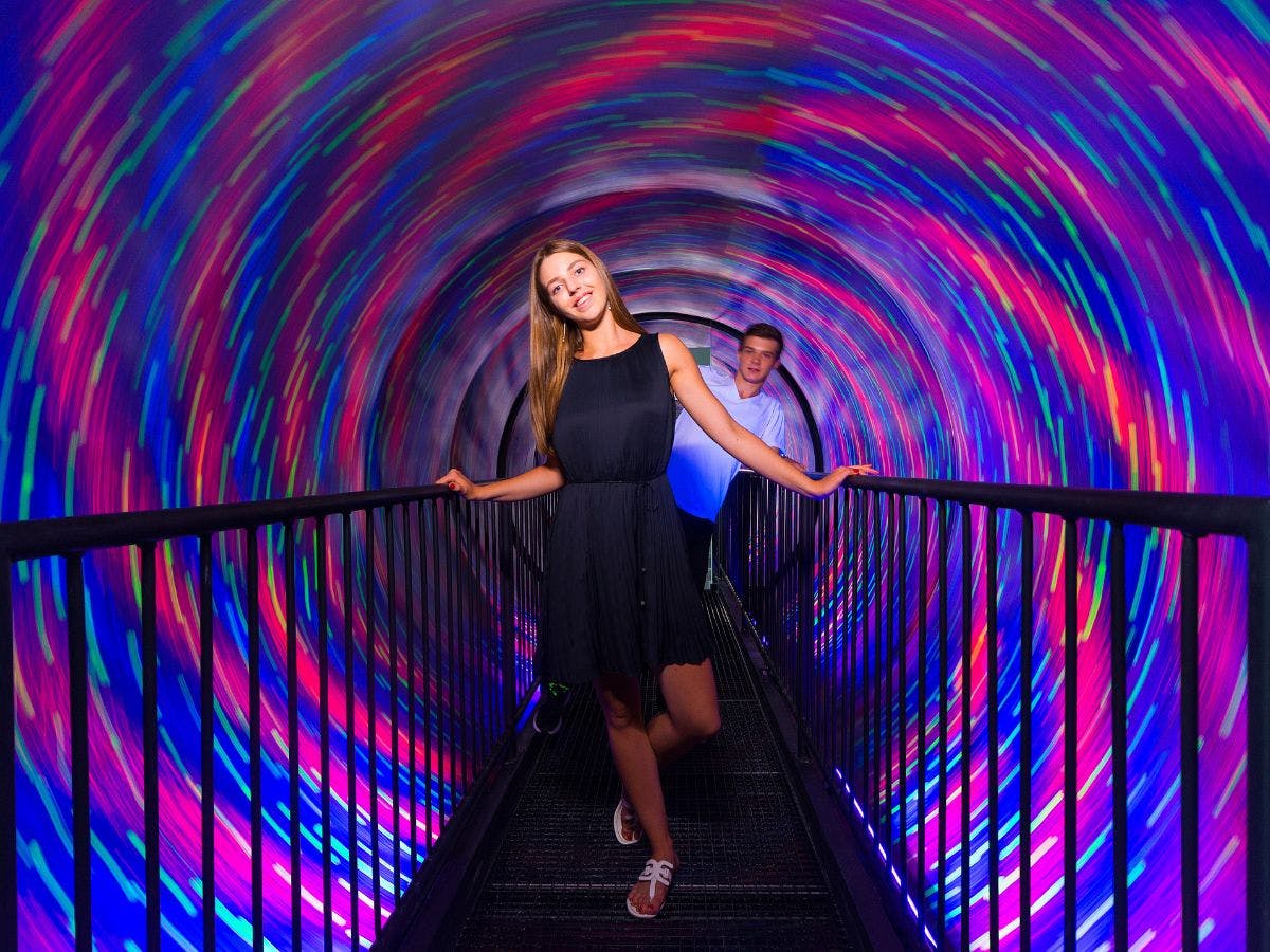 Zwei Menschen gehen durch einen rotierenden Tunnel mit bunten, wirbelnden LED-Lichtern. Die Frau ist vorne, beide lächeln.