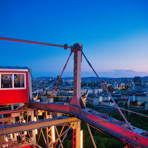 Röd pariserhjulshytt med passagerare, med utsikt över en stadsbild i skymningen med byggnader och avlägsna kullar under en klar himmel.