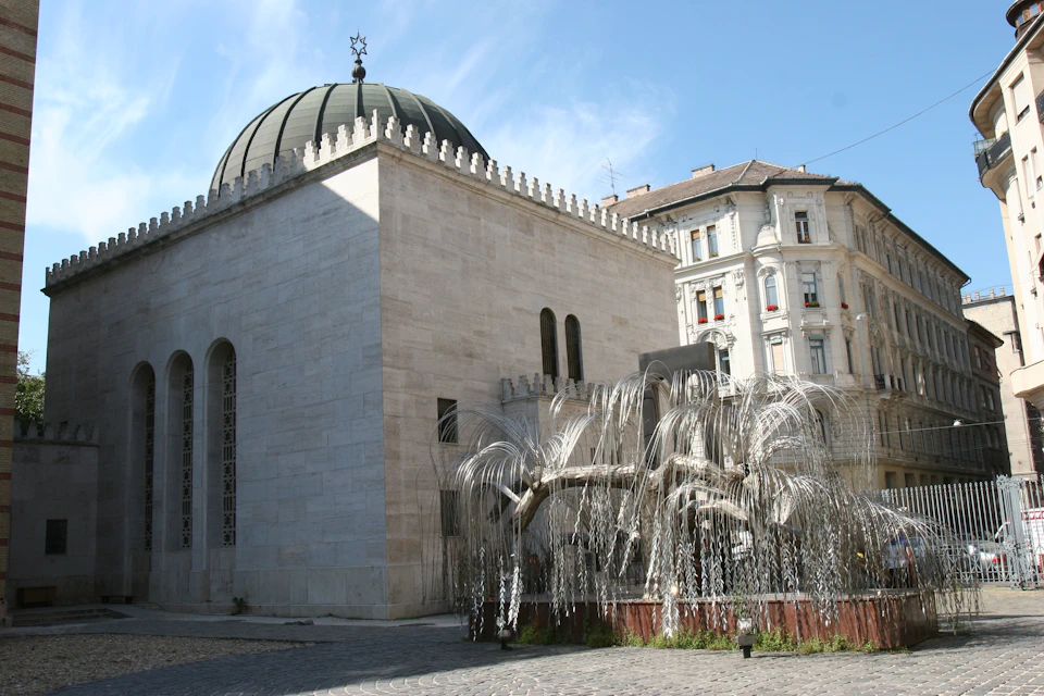 A domed building with arched windows and a metal tree sculpture outside, next to a multi-story residential building.
