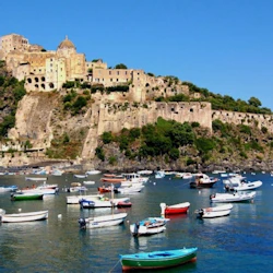 Befäst ö med historiska byggnader på en stenig kulle, omgiven av förankrade båtar och lugnt blått hav.