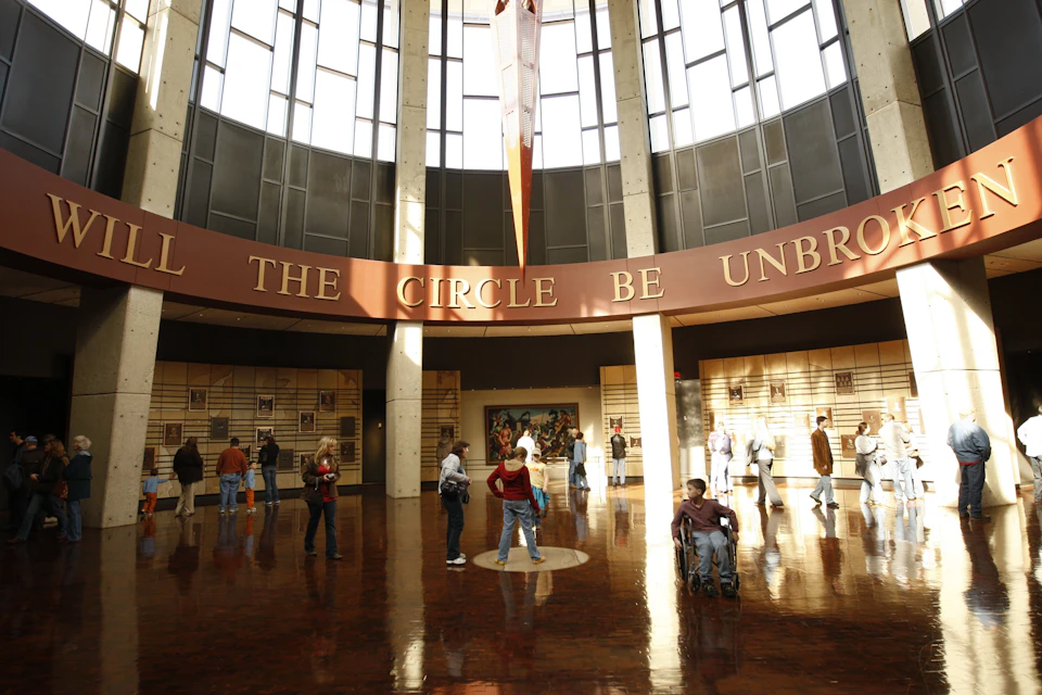People standing and walking inside a spacious hall with high windows. The walls display text: "THE CIRCLE BE UNBROKEN."