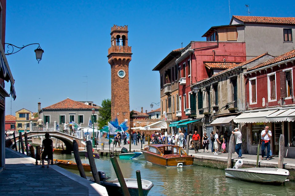 A canal with boats, a crowd by the waterfront, colorful buildings, and a tall brick clock tower under a clear blue sky.