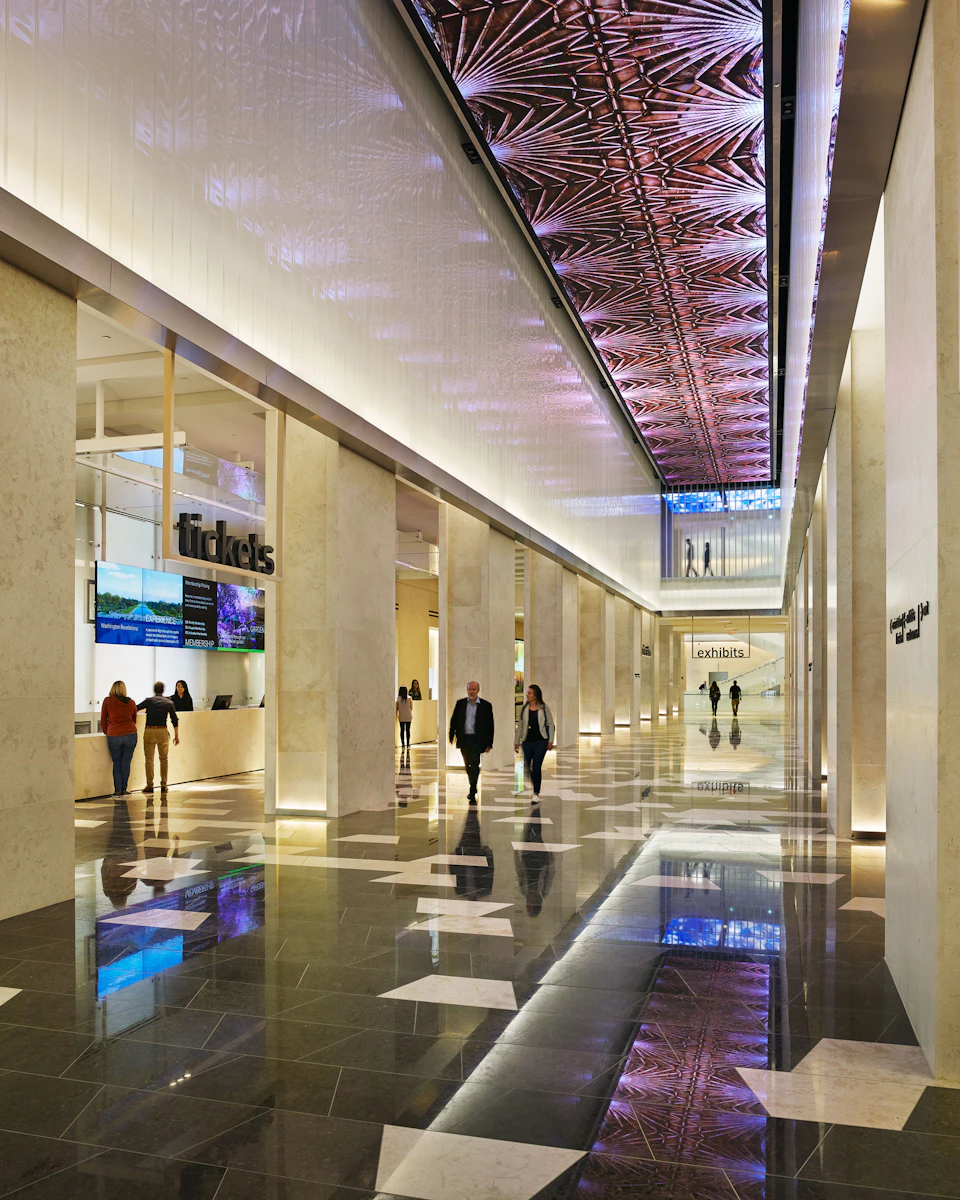 Spacious modern lobby with illuminated ceiling design, ticket counters on the left, people walking through, and reflective floors.