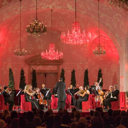 Orkester som uppträder i en elegant inredd sal med röd belysning, ljuskronor och en publik som tittar på.