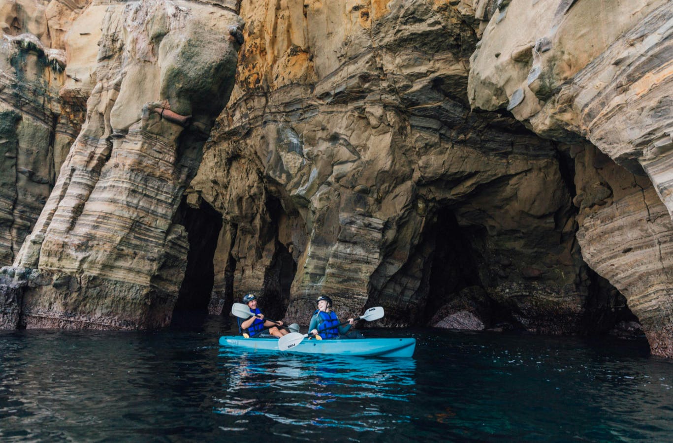 Due persone con giubbotti di salvataggio blu remano in kayak all'interno di una grotta rocciosa con formazioni di pietra a strati sopra l'acqua limpida.