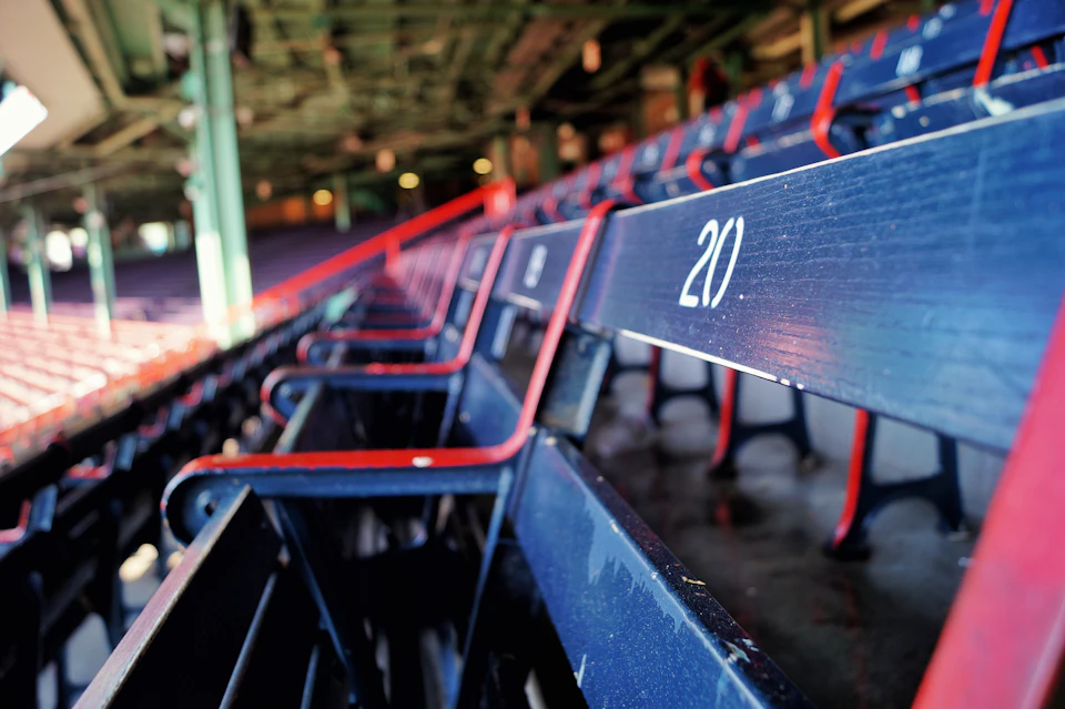 Close-up of empty, numbered blue stadium seats with red armrests under a green structure. Seat 20 is prominently shown.
