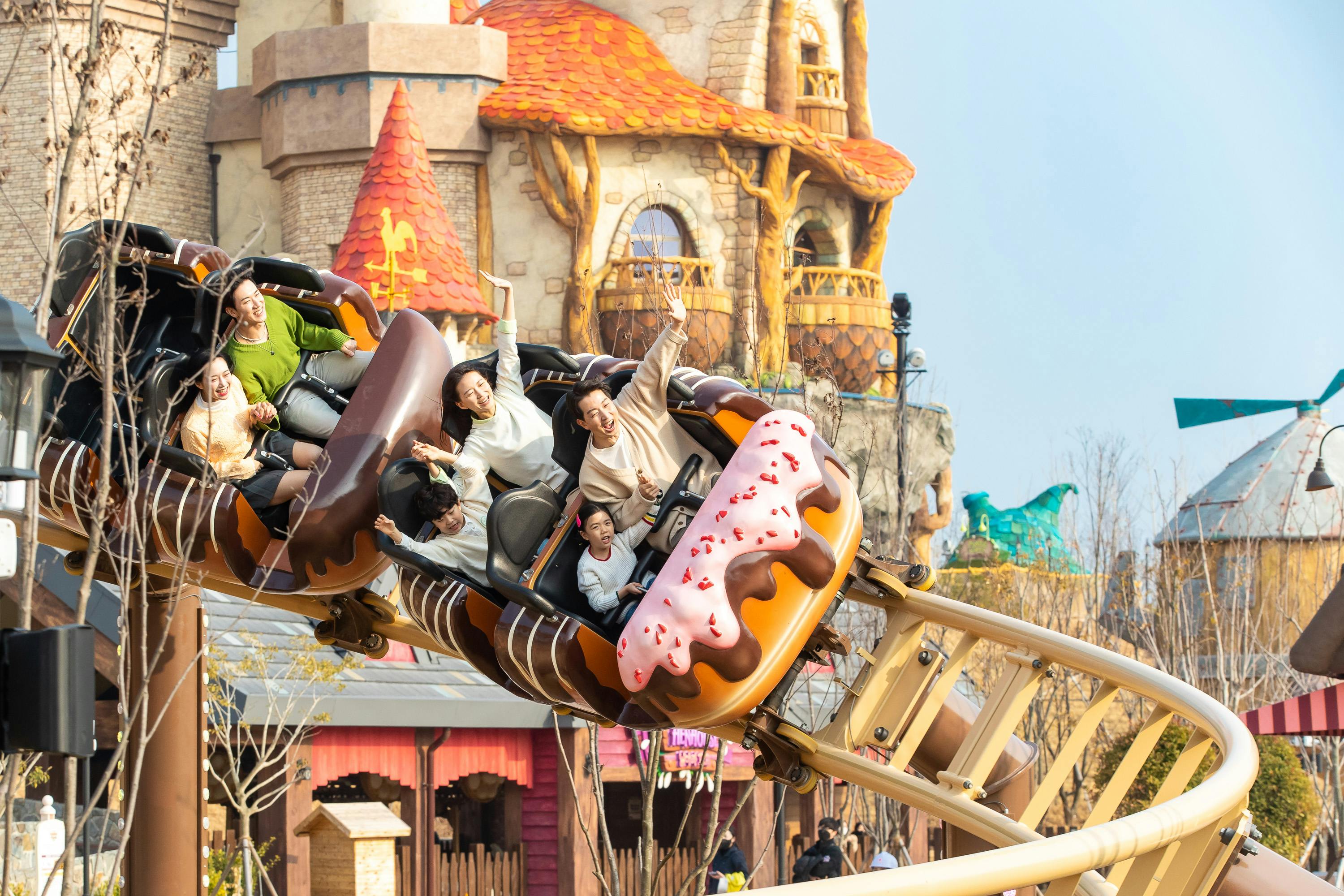 People enjoying a themed rollercoaster with donut-shaped cars in an amusement park with decorative buildings in the background.