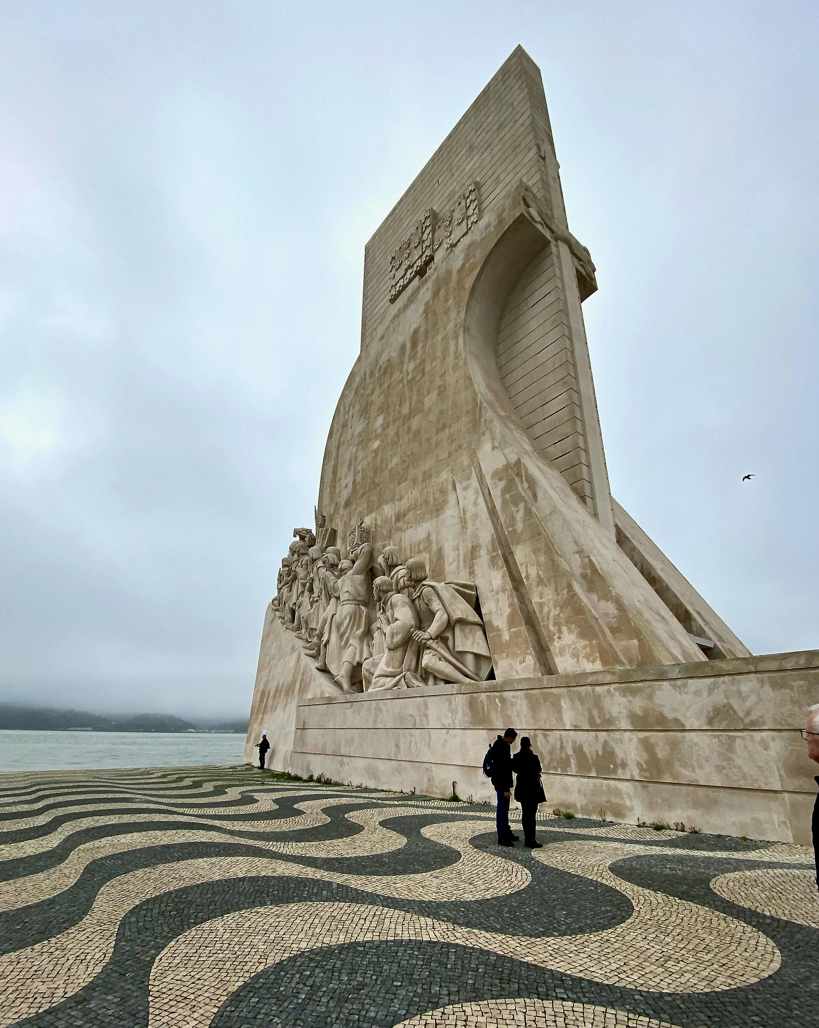 Högt stenmonument med detaljerade skulpturer på sidan. Människor står i närheten på en svartvit vågmönstrad trottoar. Molnig himmel.