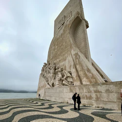 Högt stenmonument med detaljerade skulpturer på sidan. Människor står i närheten på en svartvit vågmönstrad trottoar. Molnig himmel.