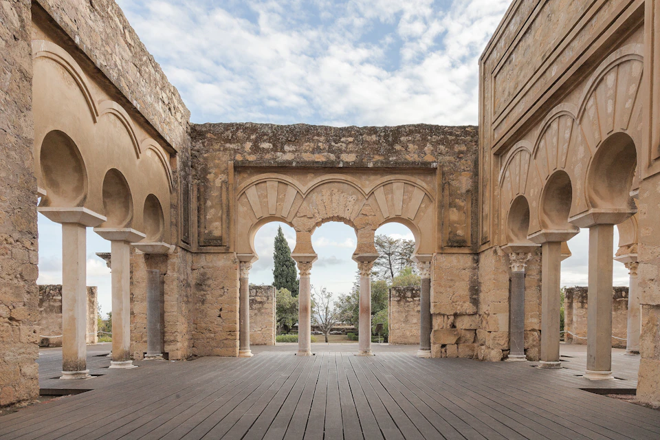 A partially-ruined stone structure with arches and columns, an open sky above, and trees visible through the arches.