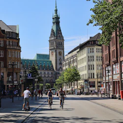 Människor som cyklar och en skoter på en stadsgata, med historisk arkitektur och ett högt klocktorn i bakgrunden.