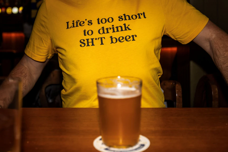A yellow shirt with text saying "Life's too short to drink SH*T beer" and a glass of beer on a table in the foreground.