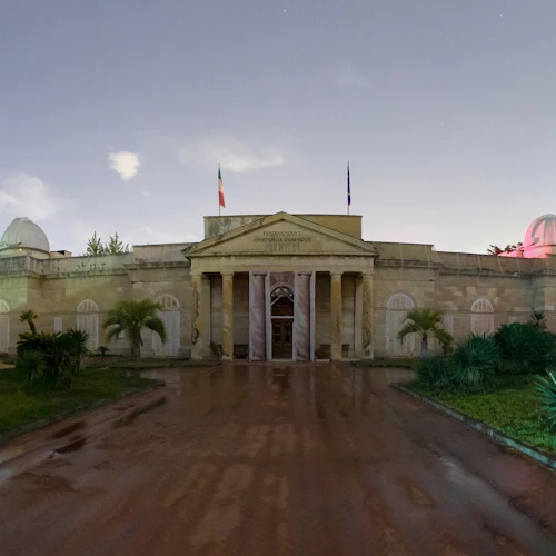 Neoklassisk observatoriebyggnad i skymningen, med pelare, kupoler, flaggor och omgivande grönska mot en molnig himmel.