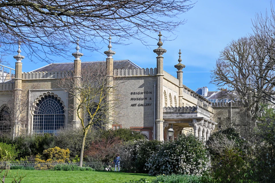 Historic building labeled "Brighton Museum & Art Gallery," surrounded by trees and bushes, under a clear blue sky.