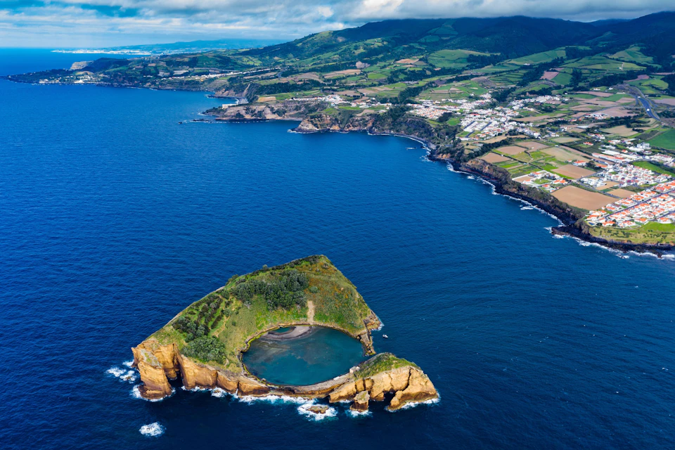 Aerial view of a coastal landscape with an island featuring a circular lagoon, surrounded by cliffs and bordered by farmland and villages.