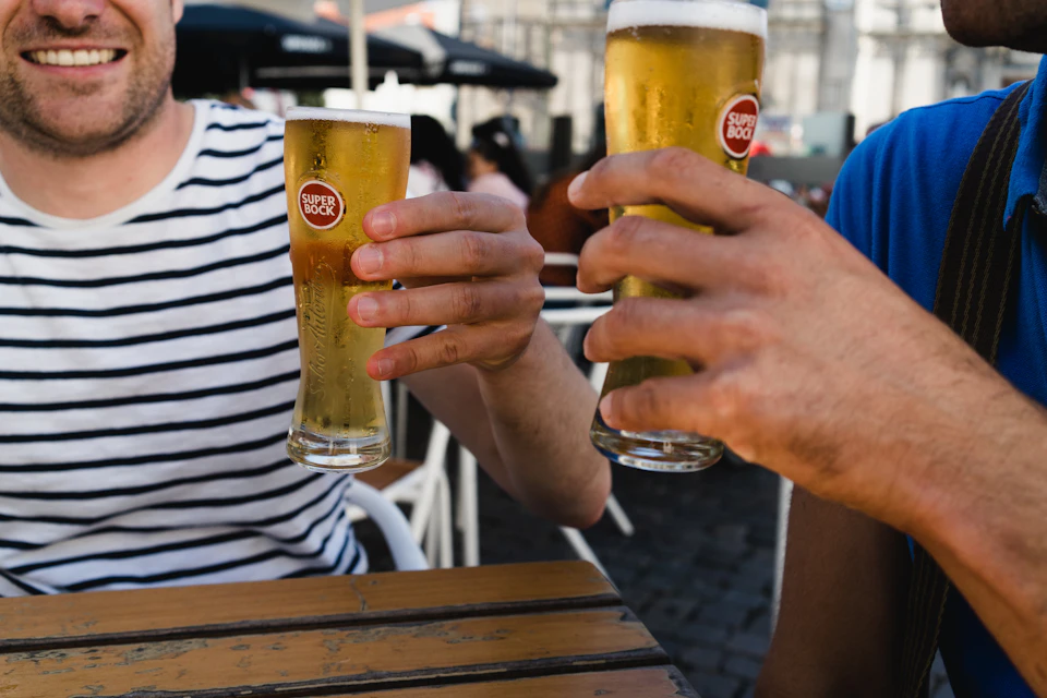 Two people clinking glasses of Super Bock beer at an outdoor café, one in a striped shirt and one in a blue shirt.