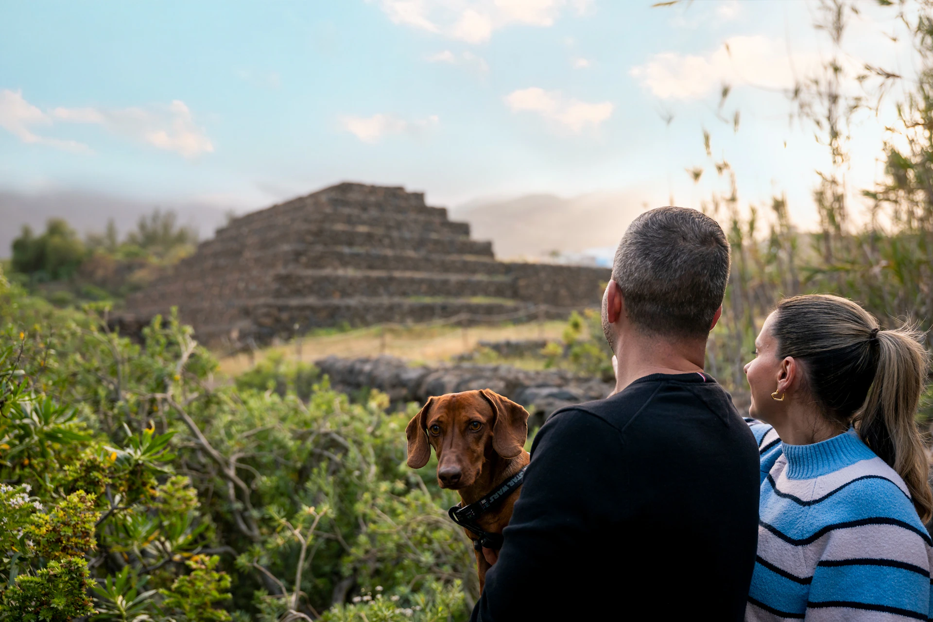 A man, a woman, and a dog look at a stepped stone structure in an outdoor setting with a cloudy sky.