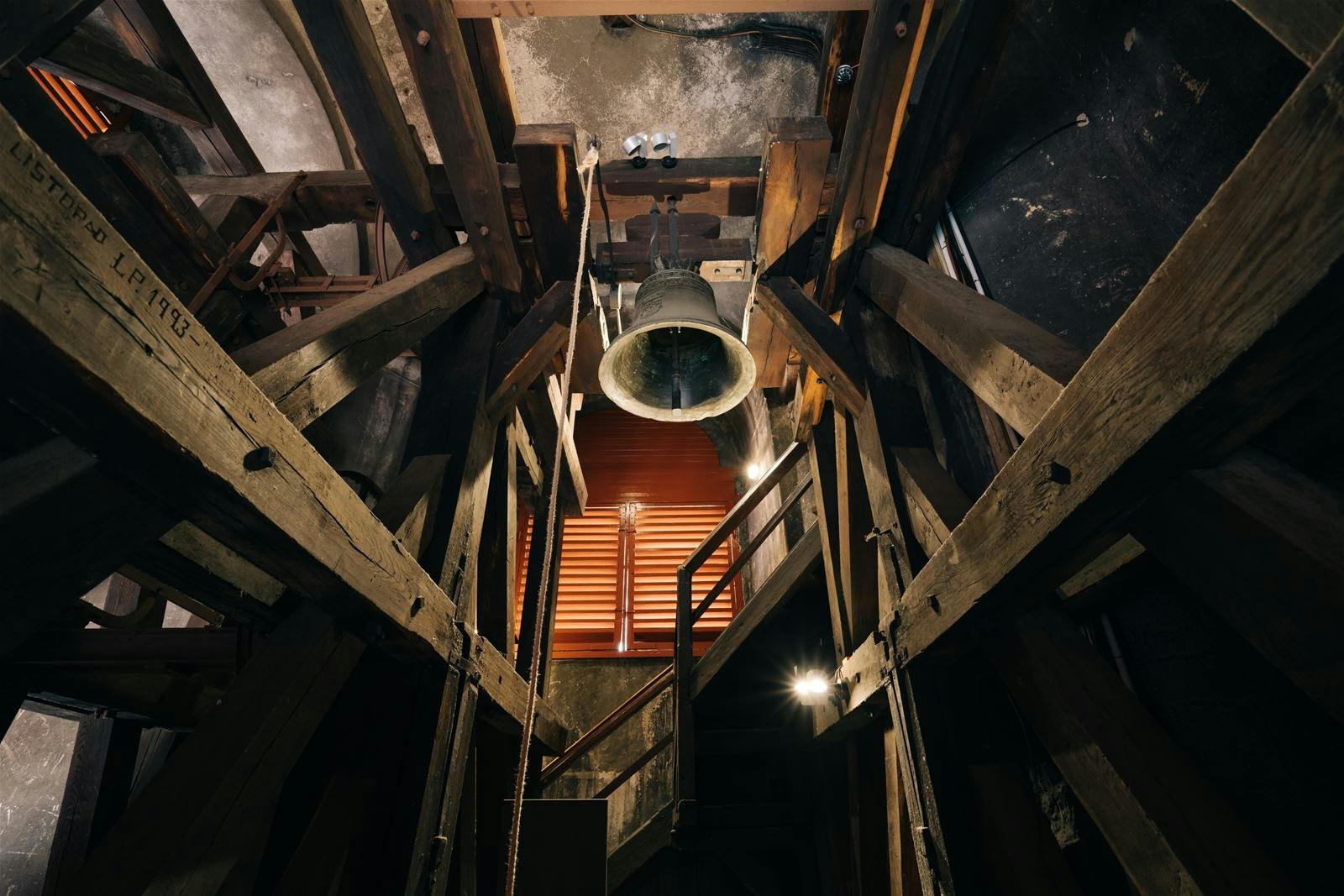 Gammelt klokketårnsinteriør med stor klokke, træbjælker og trapper. Lys fra et lille vindue oplyser rummet.