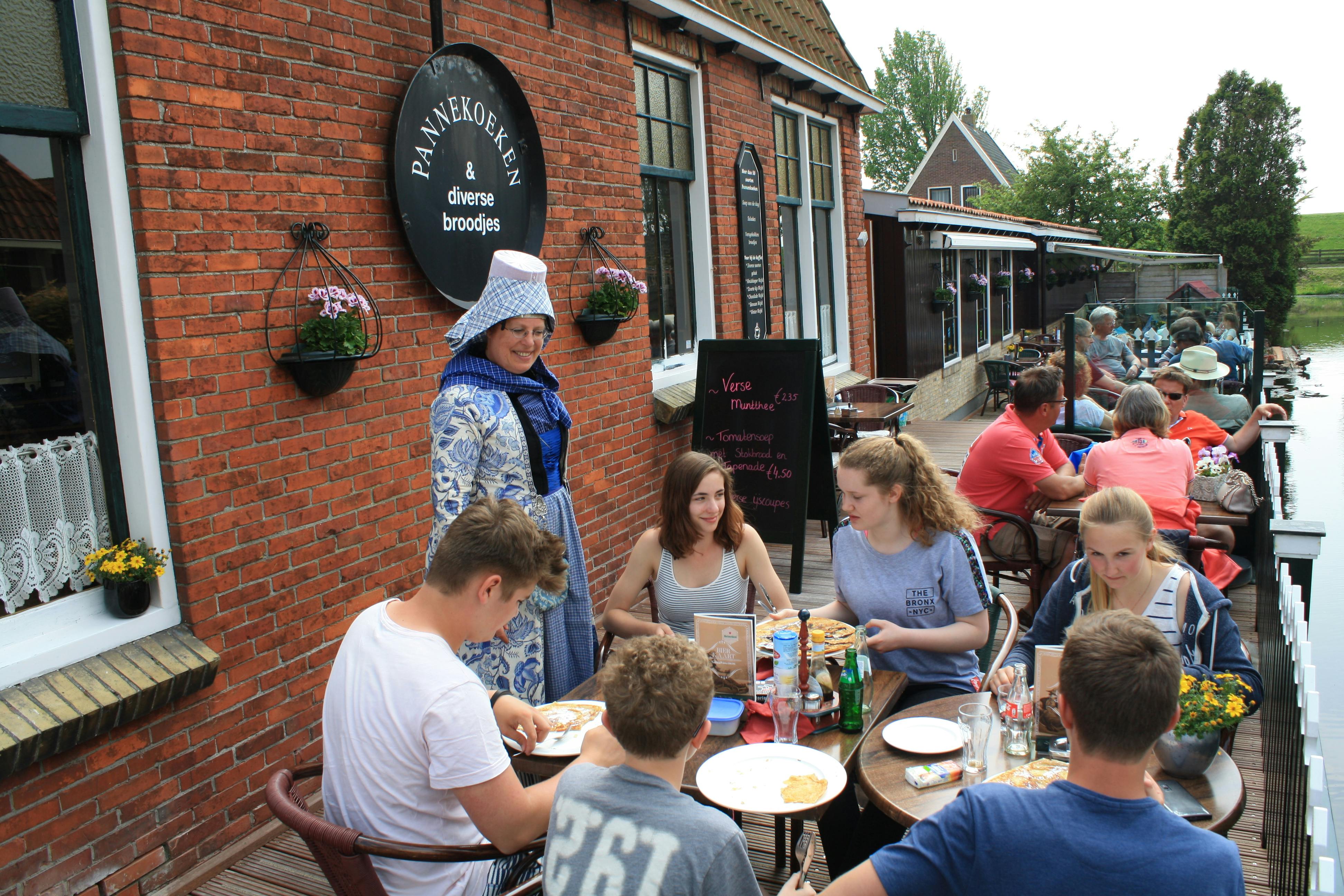 '판넨코켄 (Pannenkoeken)' 이라는 간판과 네덜란드 전통 의상을 입은 서버가 있는 카페에서 야외 식사를 즐기는 사람들.