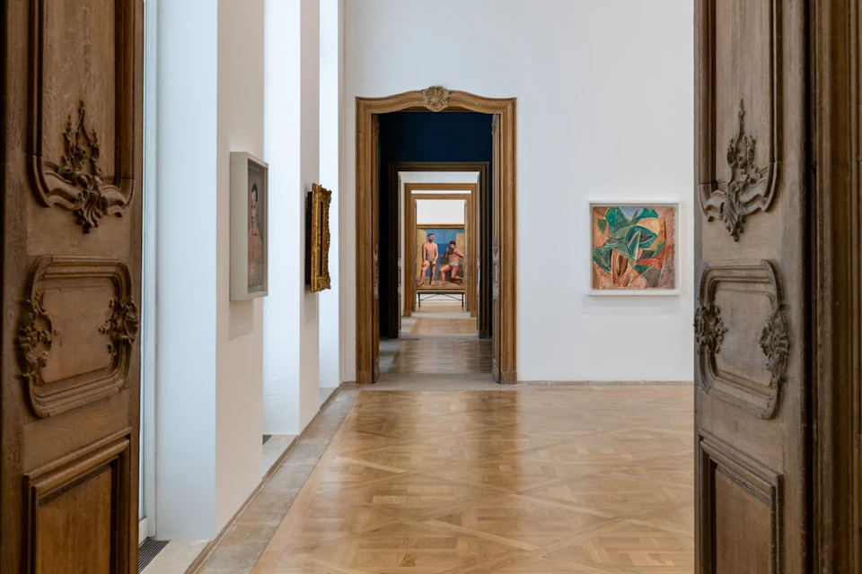 A gallery hallway with wooden floors, white walls, framed artwork, and a series of open doorways creating a visual depth.