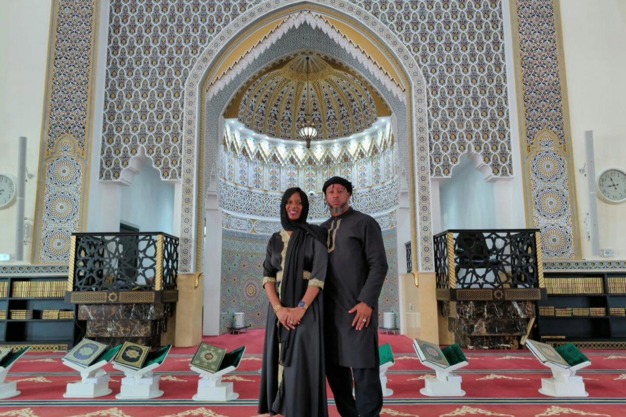 Two people stand in traditional attire inside an ornate mosque with intricate patterns and a decorative mihrab.