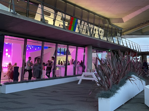 Ludzie mieszający się wewnątrz budynku z oznakowaniem „BAUHAUS”, oglądani przez duże okna z fioletowym oświetleniem. Rośliny na świeżym powietrzu i biała ławka.