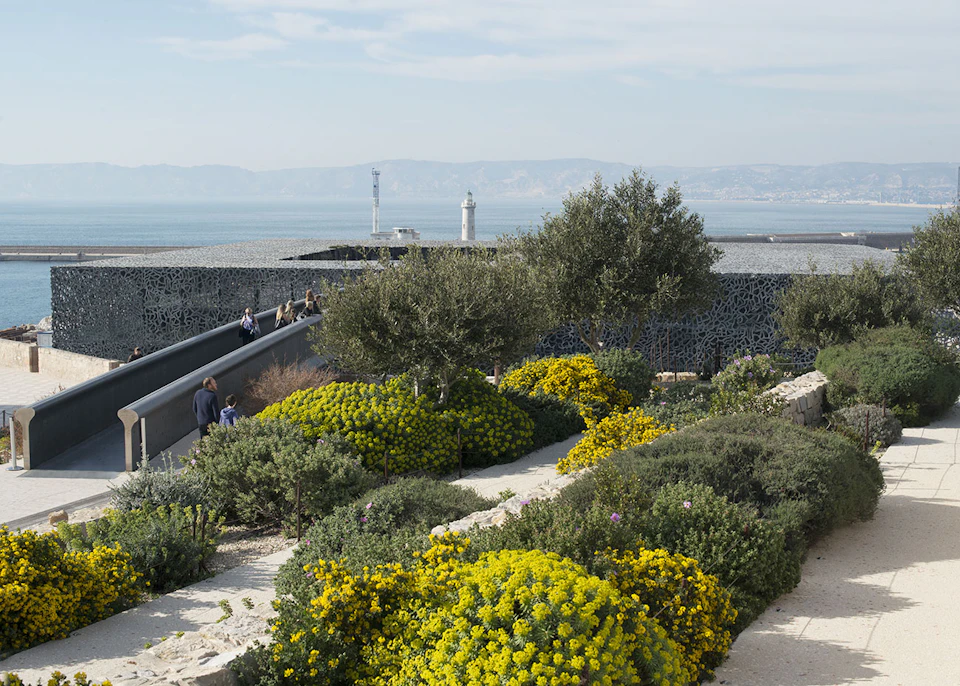 People walking on a path through a lush garden with yellow flowers, overlooking the sea and lighthouses in the background.