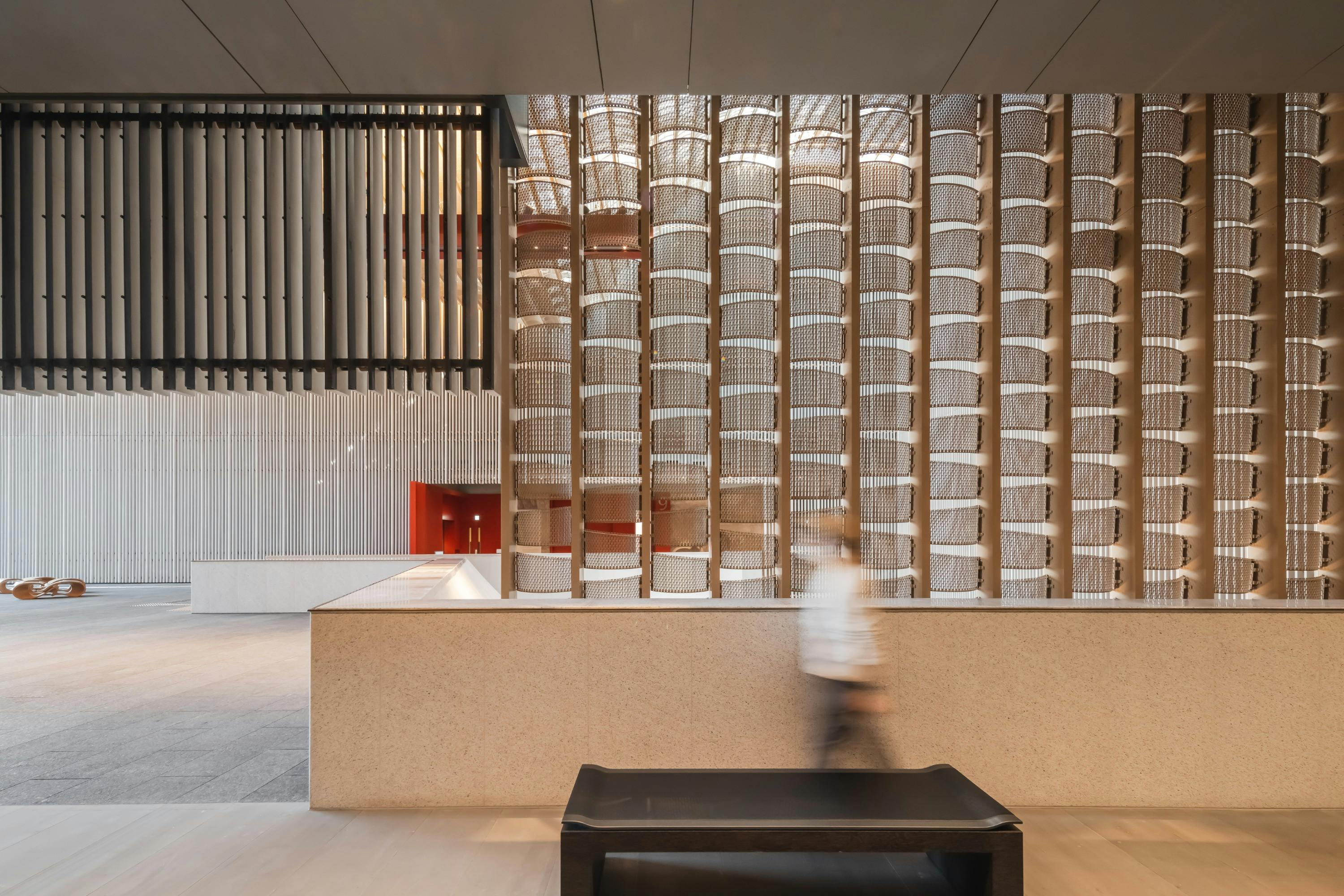 Modern interior with tall, textured panels, a red accent wall, and a blurred person walking near a bench.