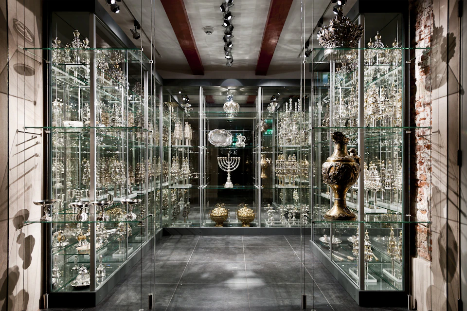 A glass-enclosed display room with shelves filled with various silver and gold artifacts, including menorahs and ornate vases.