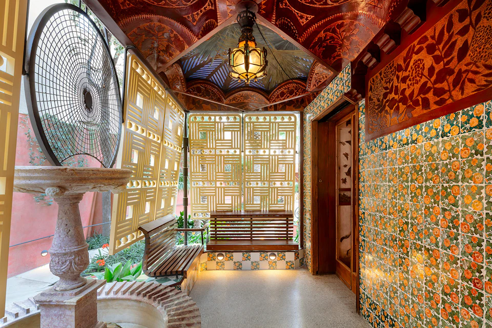 Decorative indoor space with tiled walls, a bench, intricate ceiling, fan, and a lattice window, illuminated by an ornate light.